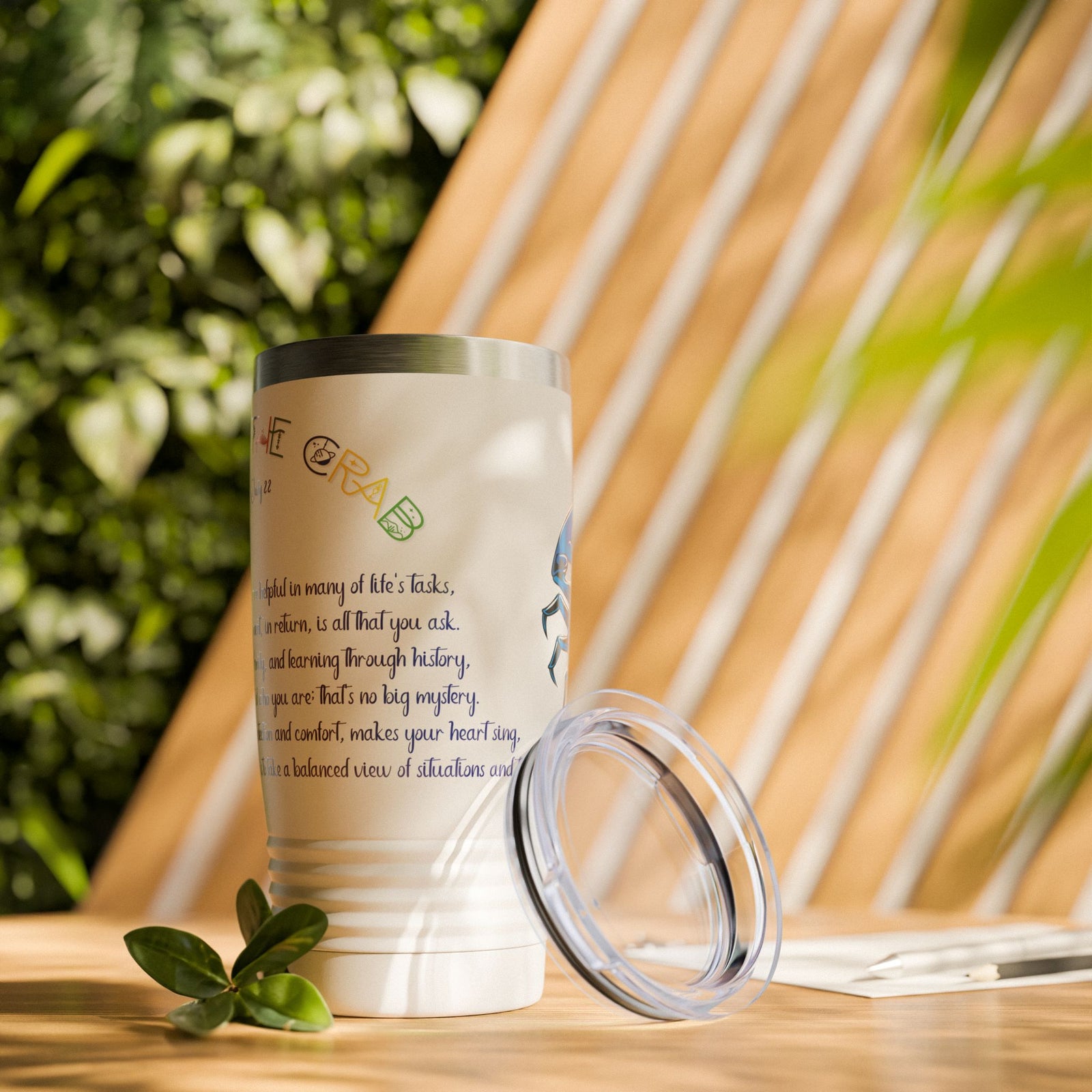 White tumbler with text and lid on a wooden surface with plants in the background