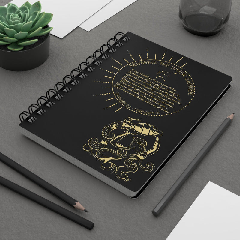 Black notebook with gold designs on a gray surface with pencils and a plant.