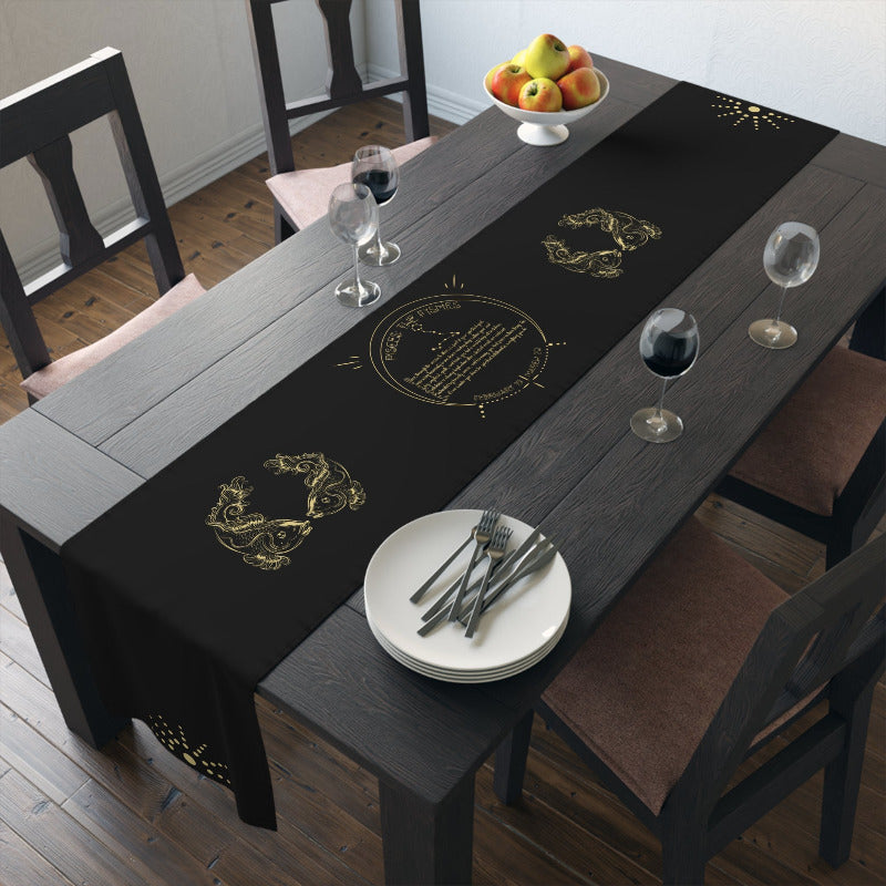 Dining table with black table runner with acrostic poem spelling out Pisces featuring gold designs, wine glasses, and a plate.